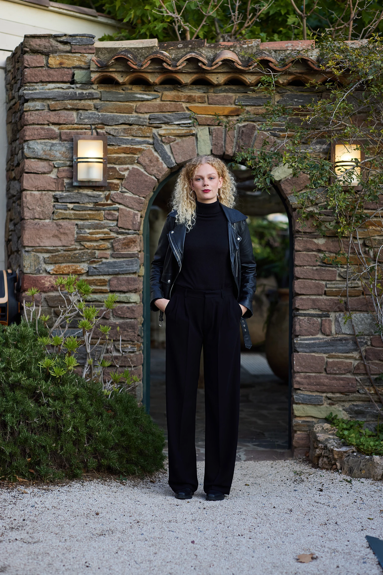 A blonde woman in black extra long pants and styled with a black leather biker jacket.