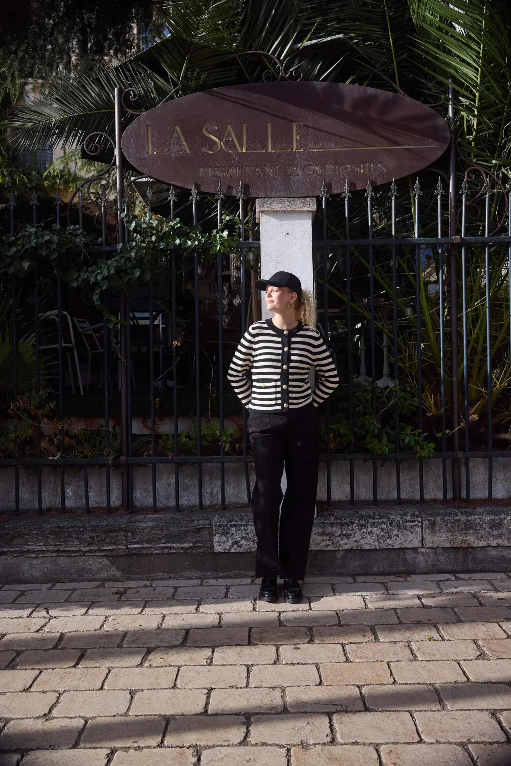 Woman wearing a striped cardigan and black orleans chinos from Gentaille, standing in front of a pillar with a french sign on top.