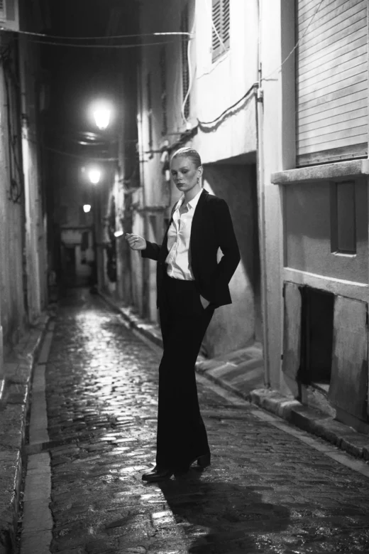 Svartvit bild på modell som har på sig en svart kostym på en liten bakgata i södra Frankrike med stämningsfullt ljus liknande Helmut Newtons klassiska bild för Yves Saint Laurent.